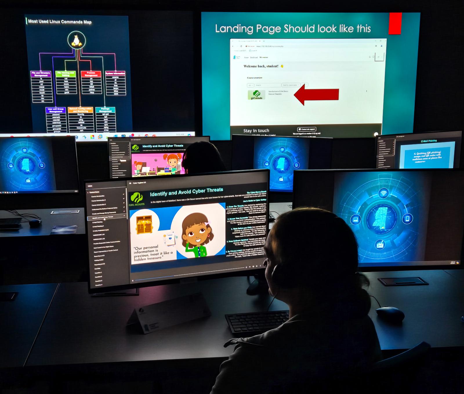 A Girl Scout learns about cybersecurity with several computer screens lit up in a dark room